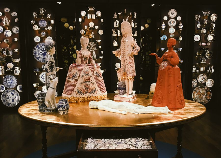 Porcelain figureines in elaborate outfits on a table. One figurine is naked and lies flat on the surface