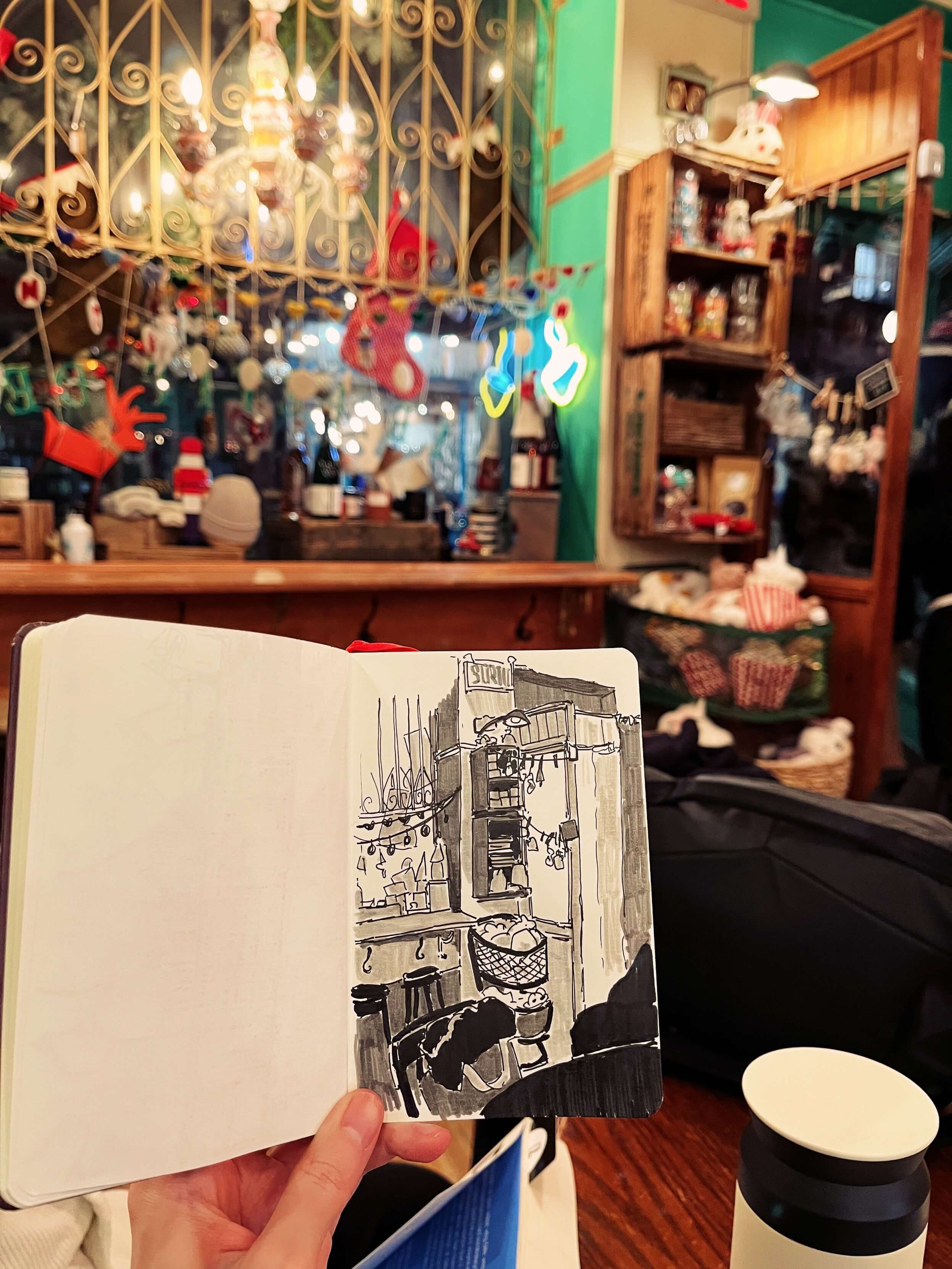 Holding up a sketchbook drawing of a cafe window. The window has a bunch of holiday decorations. Next to it are some shelves with jars and souvenirs.