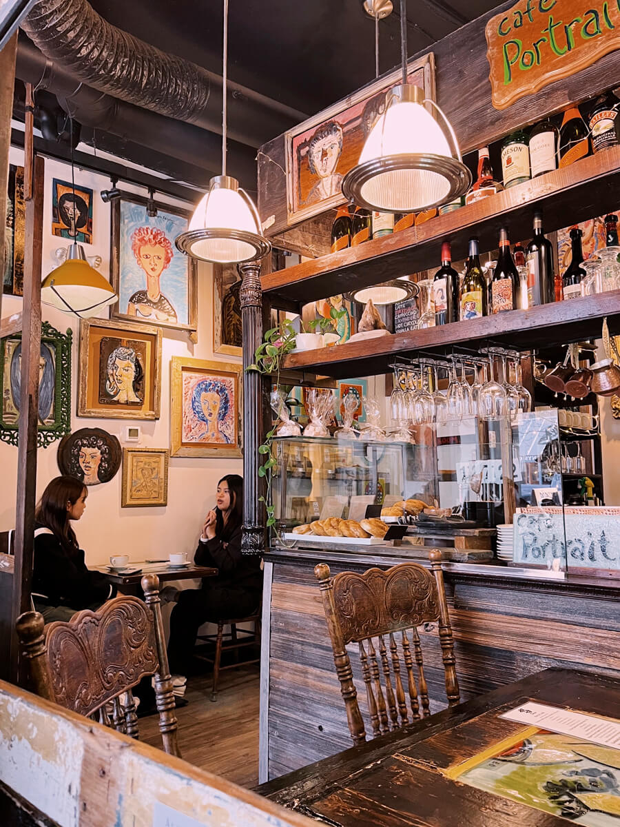 Cozy cafe interior full of worn wooden surfaces. The wall is full of portrait paintings with mismatched frames.