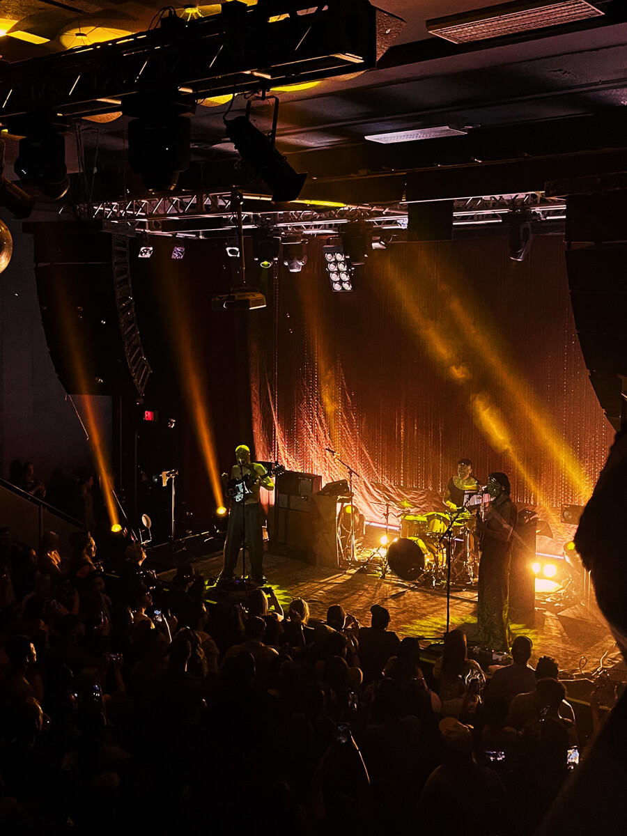 Concert photo of a guitarist, bassist, and drummer performing under yellow lights, viewed from the top right side.