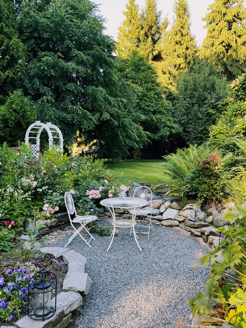 A white patio set in a yard, surrounded by flowers and trees.