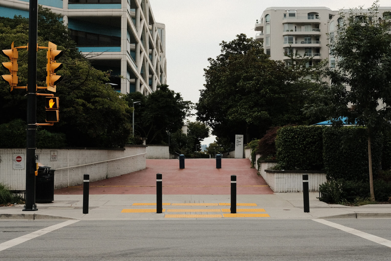 Looking across a crosswalk to the other side, where a pedestrian path leads up a slope and into a walkway between some buildings. The path is lined with trees.