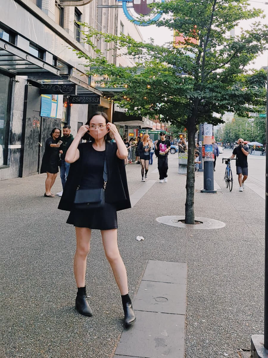 Me standing on a street, putting on a mask and wearing a black dress, jacket, and boots.