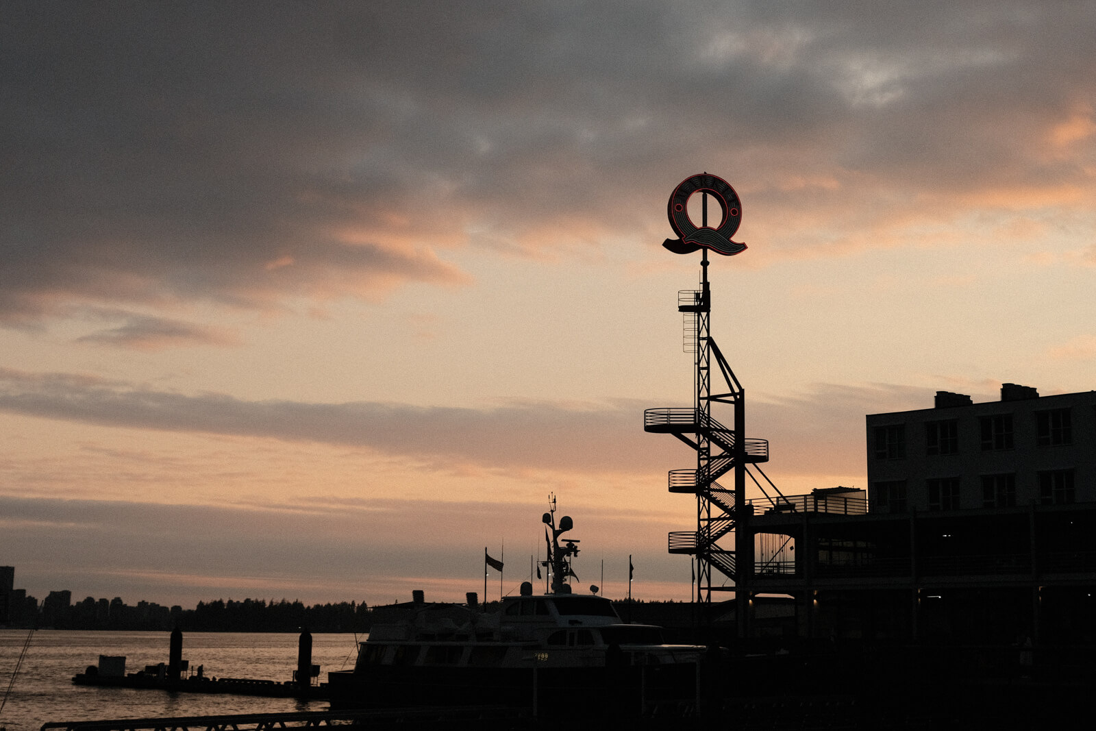 The silhouette of the 'Q' structure, framed by some small buildings. It sits on the edge of a pier. It's sunset and the sky and clouds are many shades of orange, blue, and yellow.