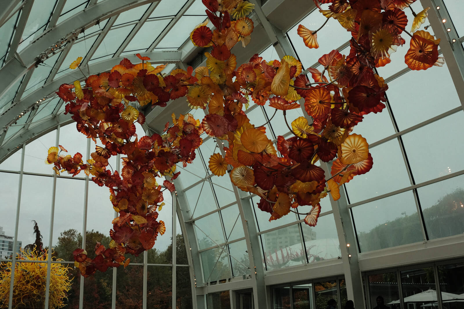 A huge glass installation suspended from the ceiling of a tall greenhouse. The installation is a winding structure of countless red, orange, and yellow flowers.