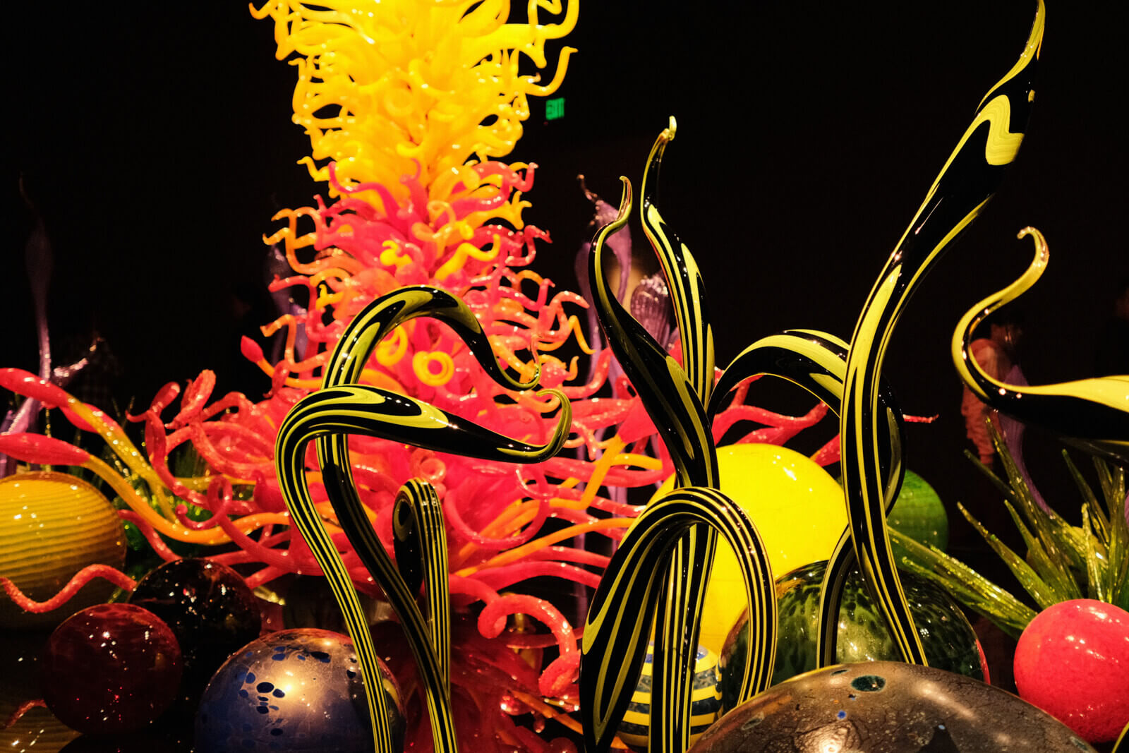 A glass sculpture installation, reminiscent of the sea floor. In the front are slim, winding tendrils in neon green and black, resembling seaweed. In the background are glass spheres of varying sizes, colours, and patterns, as well as a large column of countless tendrils.