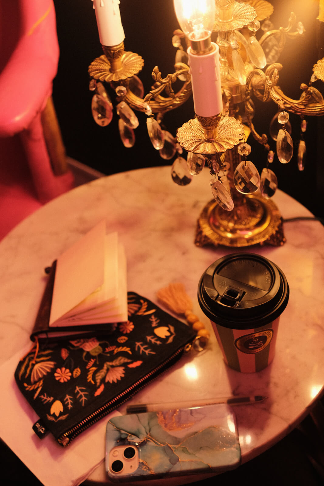 My sketchbook, pencil case, phone, and coffee scattered about on a round marble table. An ornate candle holder, which features dangling clear gems, sits next to them.