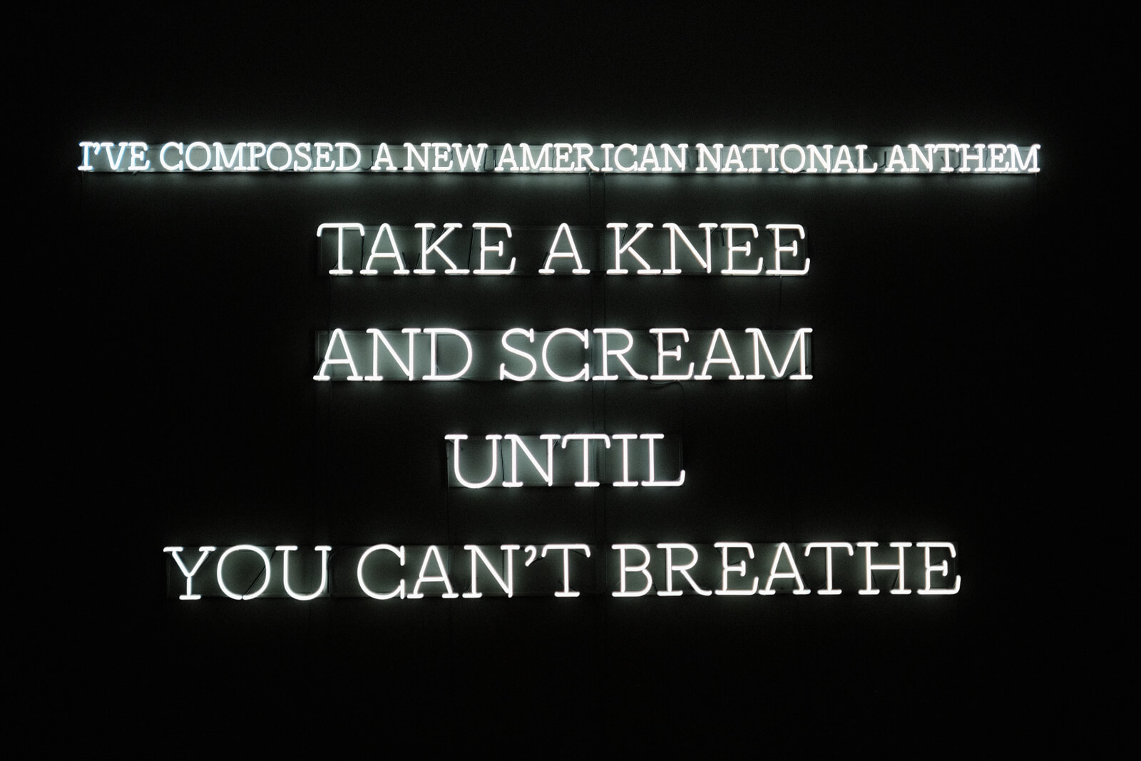 An installation of text in the form of white neon all caps text on black that reads, 'I've composed a new American National anthem. Take a knee and scream until you can't breathe.'