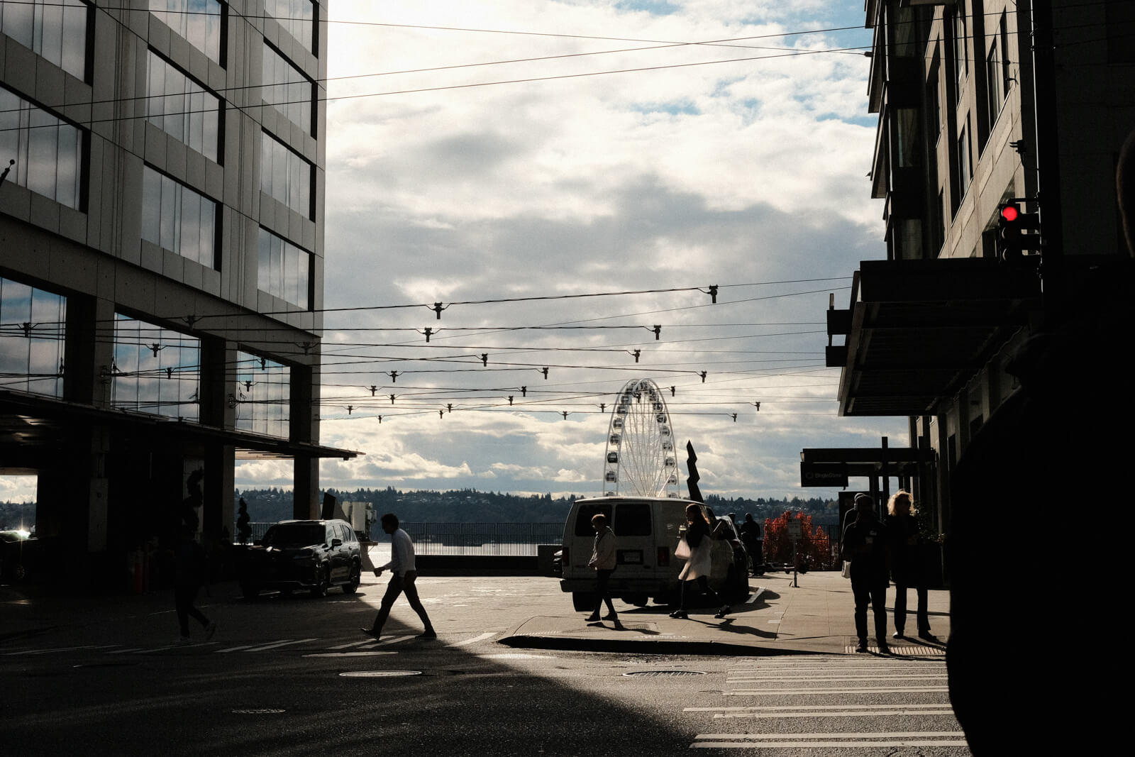 A figure crosses the street and into the shadow cast by a tall building. In the background, a ferris wheel is visible.