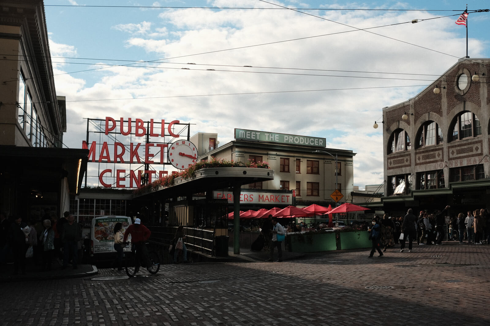 The Public Market Center sign. Many people mill about in front of the market under the sun and clouds.