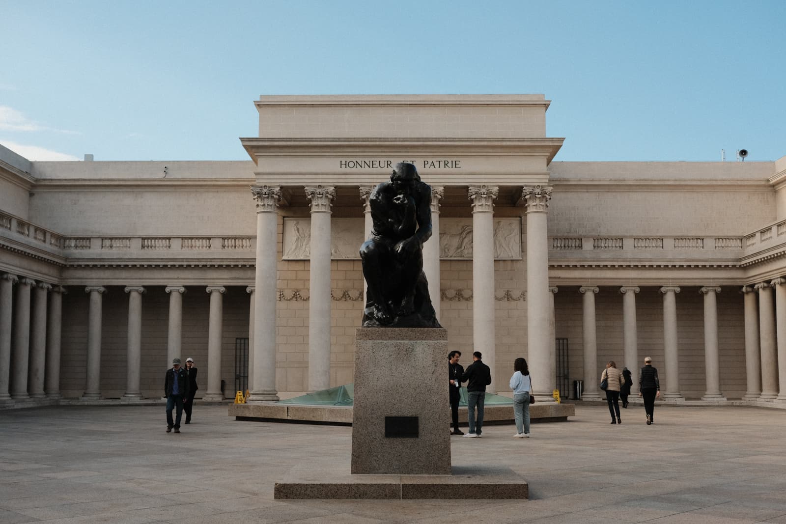 A Rodin sculpture in the outdoor plaza of a museum, which is a big fancy looking building lined with tall columns, like in the style of the Pantheon.