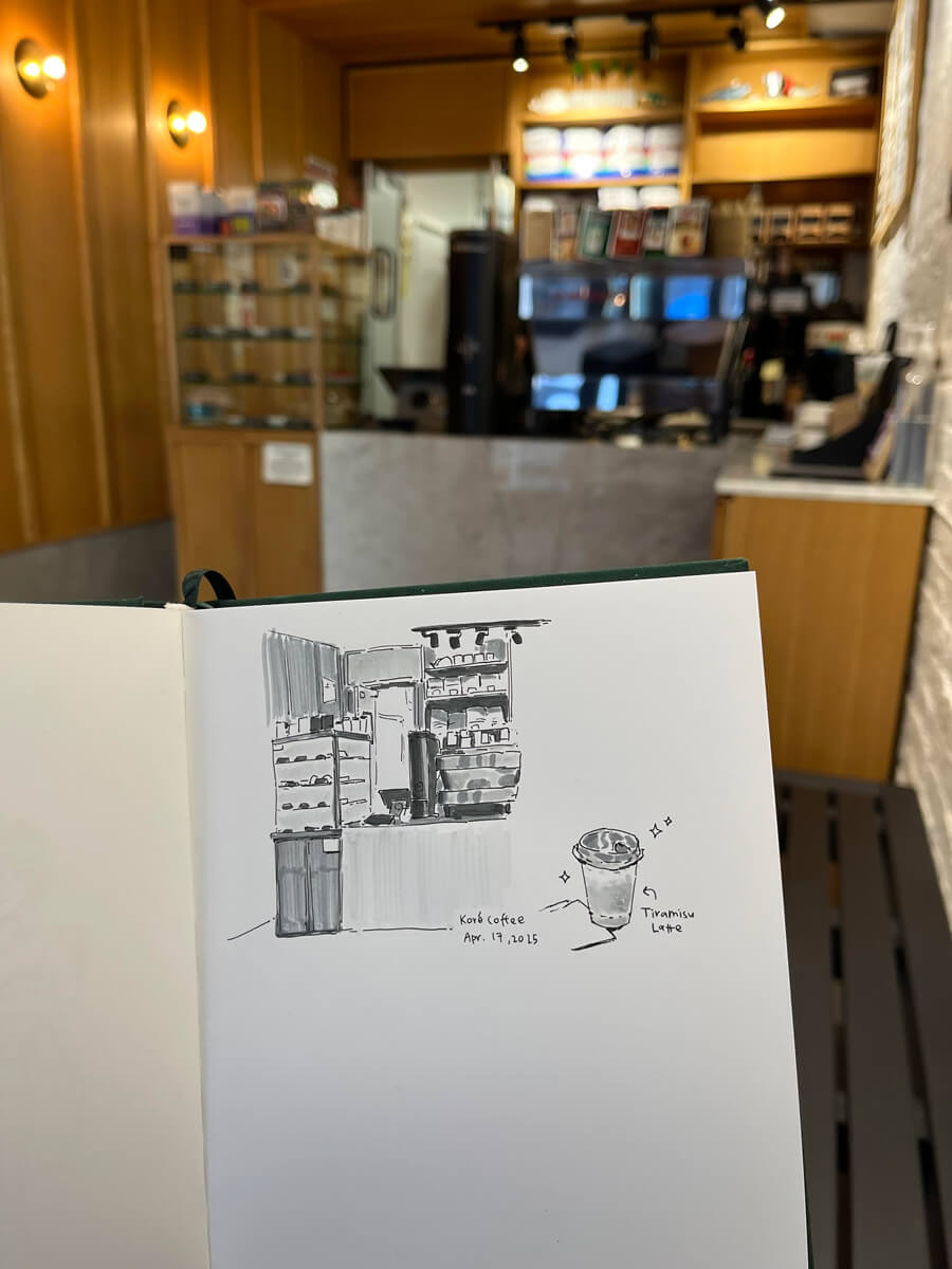 Ink sketch of the counter of a cafe and an iced drink labeled ‘tiramisu latte’