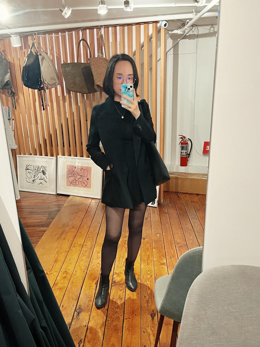 Mirror outfit photo in a store. I’m wearing an oversized blazer, high-necked top, short pleated skirt, tights, and ankle boots, all in black.
