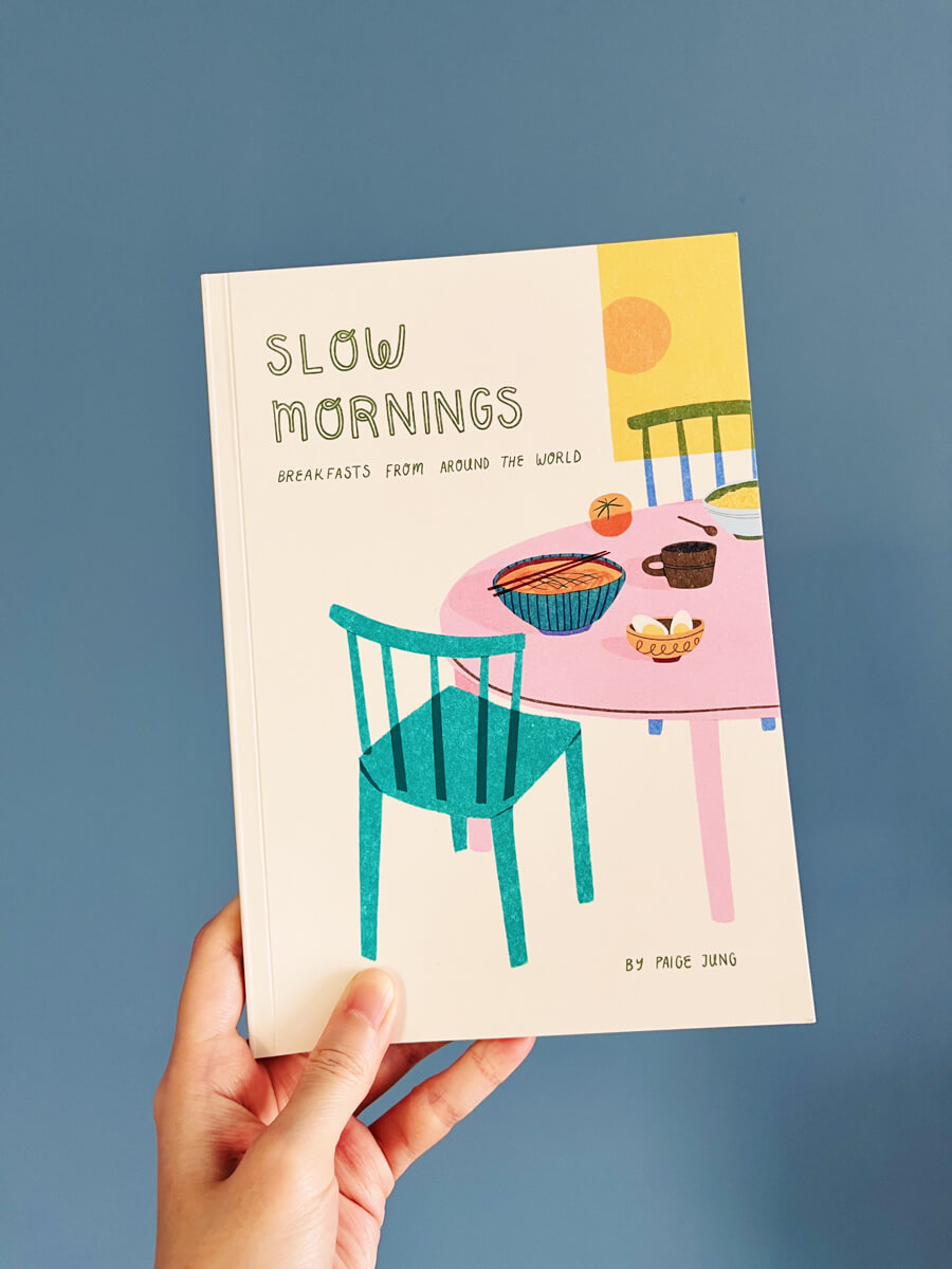 A zine of breakfasts from around the world, with a colourful cover illustration of a dining table with various foods on it.