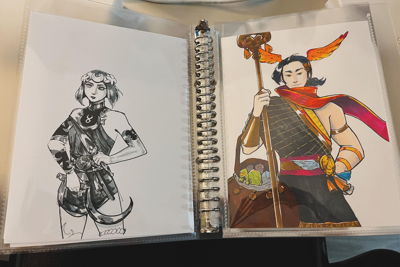 A binder laid open to show two of my Hades ink drawings stored in clear plastic sleeves.
