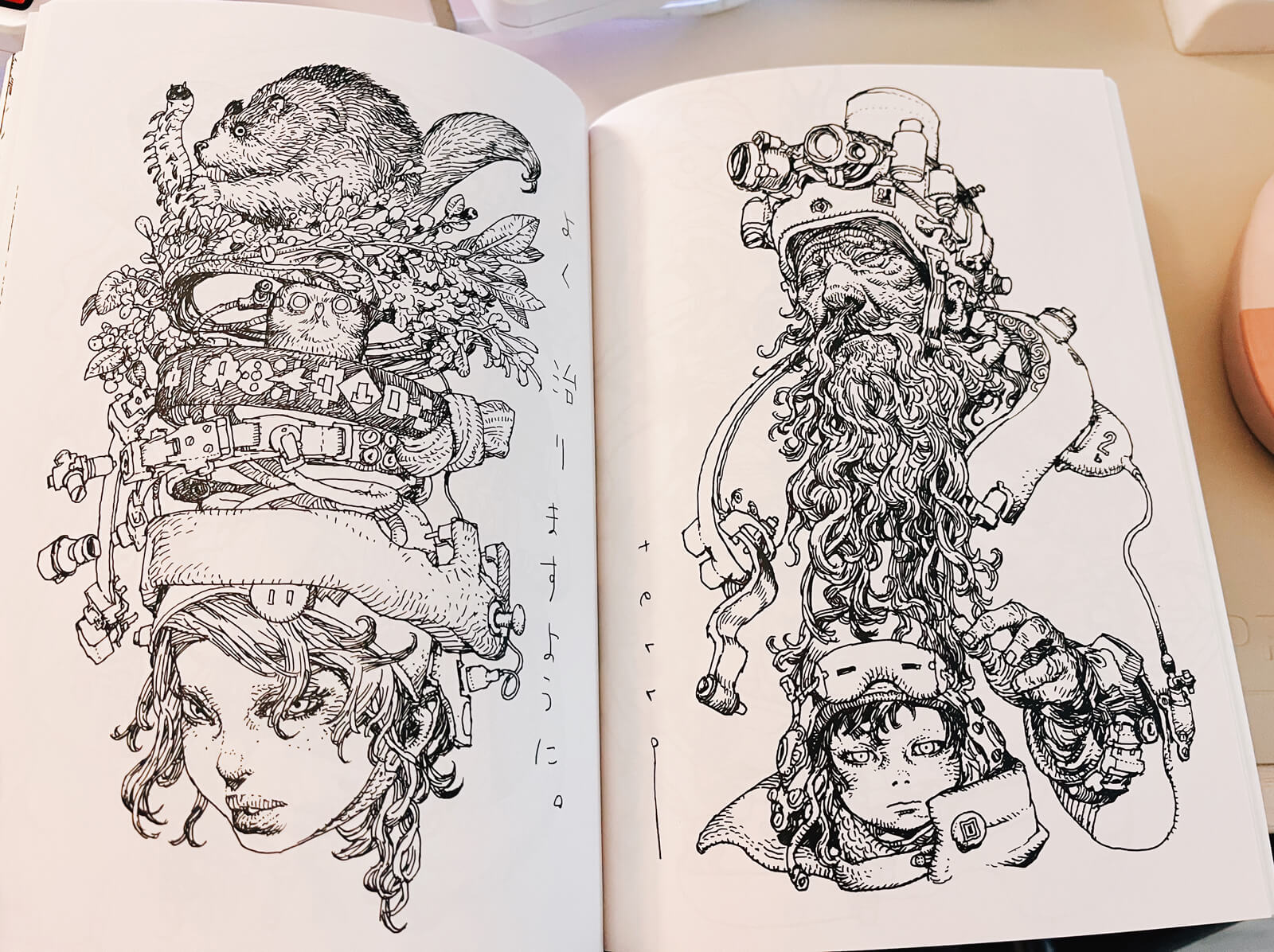 Art book open to show two ink drawings, featuring floating heads wearing intricate headpieces made of machinery, nature, and animals. It's intricately drawn, with hatching that conveys depth and texture.