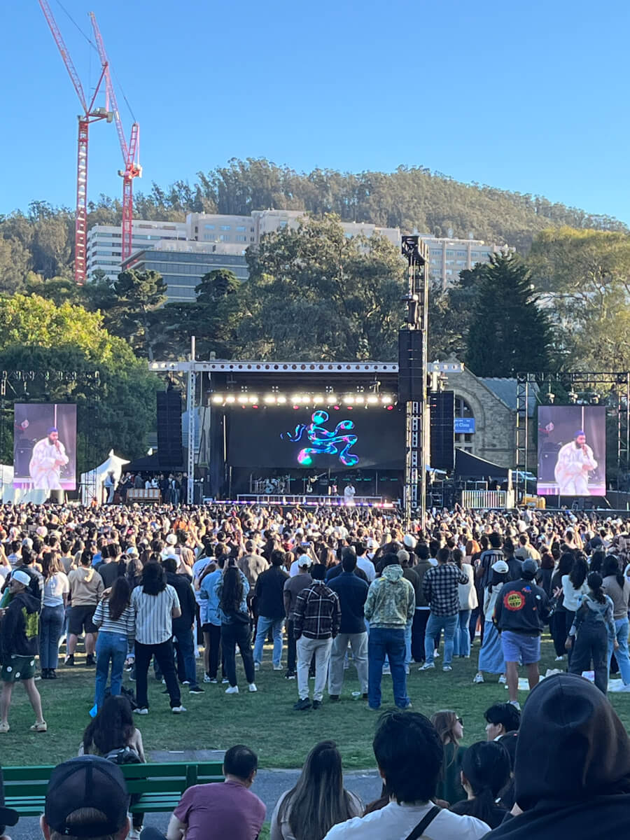 Distant view of the concert stage as Khalid performs, across a crowd of people tsanding on the grass, and closer to me, people sitting. It's bright and sunny.