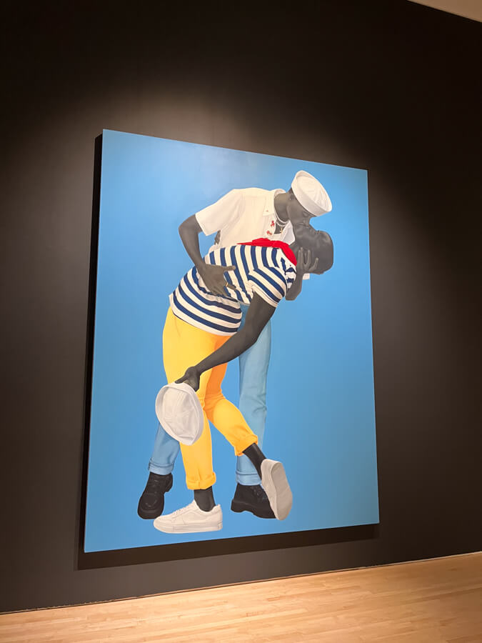 Two people in sailor hats share a kiss against a blue background. One wears a striped shirt and yellow pants, the other a white shirt and jeans.