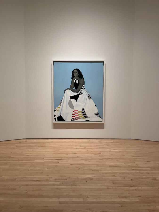 A portrait of Michelle Obama seated, resting her chin on her hand. She is wearing a long, geometric-patterned dress that features bold black, white, red, pink and yellow shapes, including stripes, triangles, and circles. The dress covers much of the lower half of the image. The background is a soft, solid light blue and her hair is styled in loose waves. Her expression is calm yet contemplative.