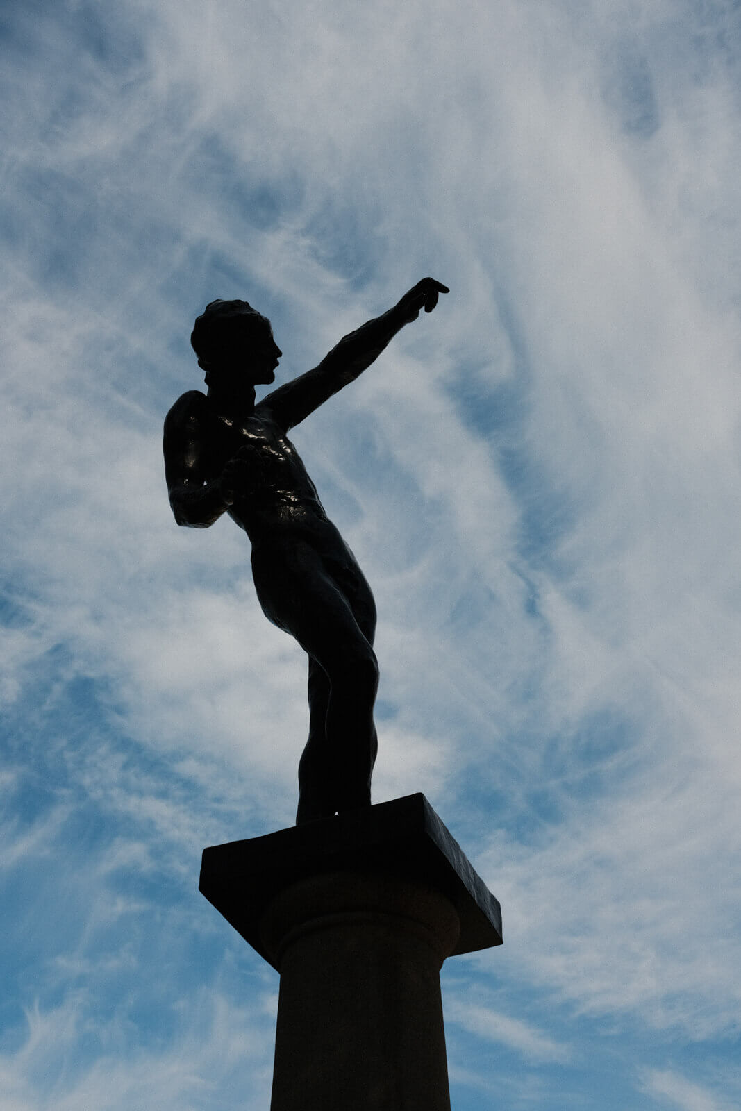 A sculpture, mostly shadowed, against a cloudy blue sky. The figure stands contrapposto with one arm reaching up.