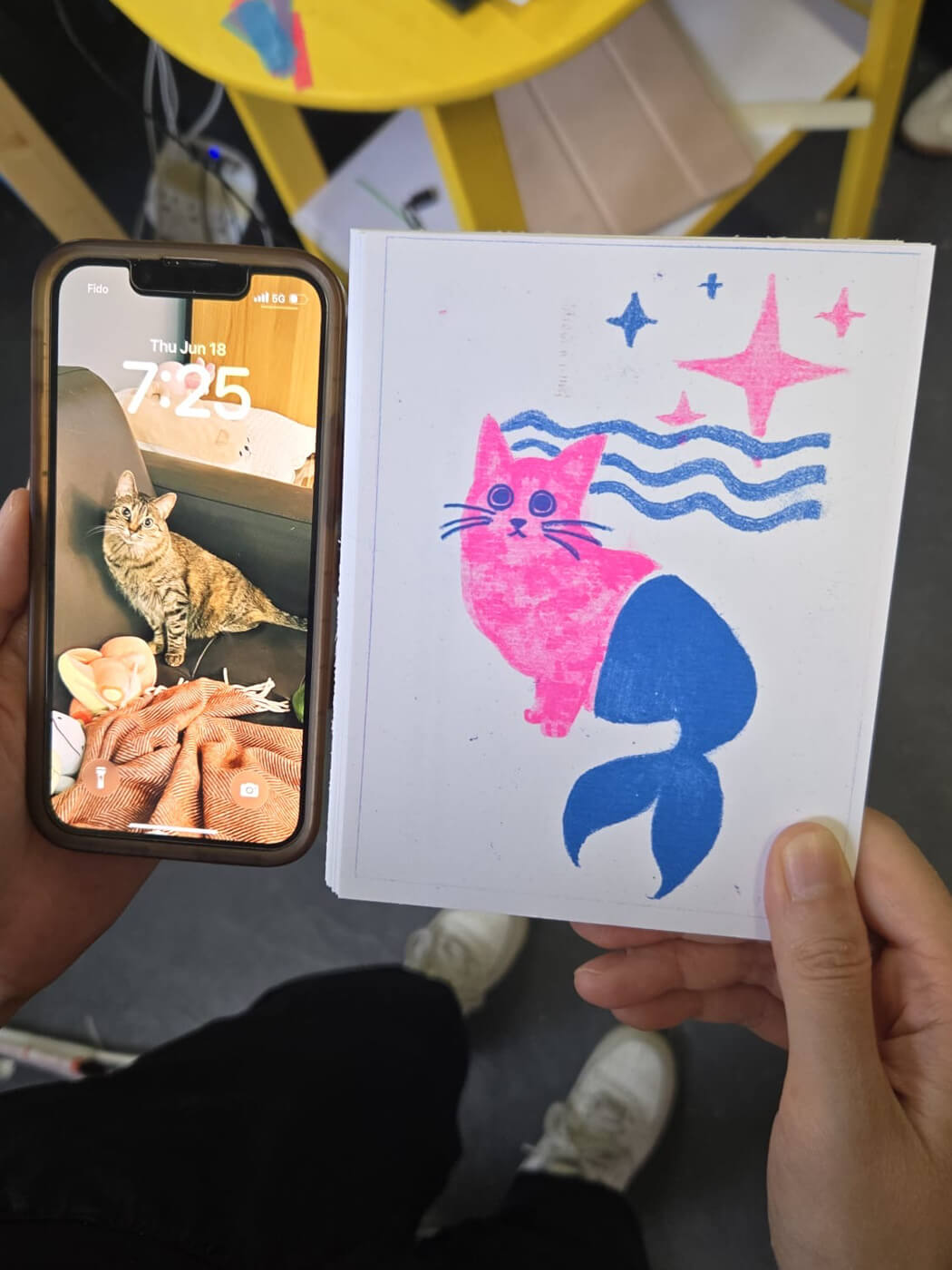Holding my riso print next to my phone. The print is a pink and blue drawing of my cat Sophie, posing like a mermaid, except her back legs are an actual mermaid tail. My phone wallpaper shows the reference photo.