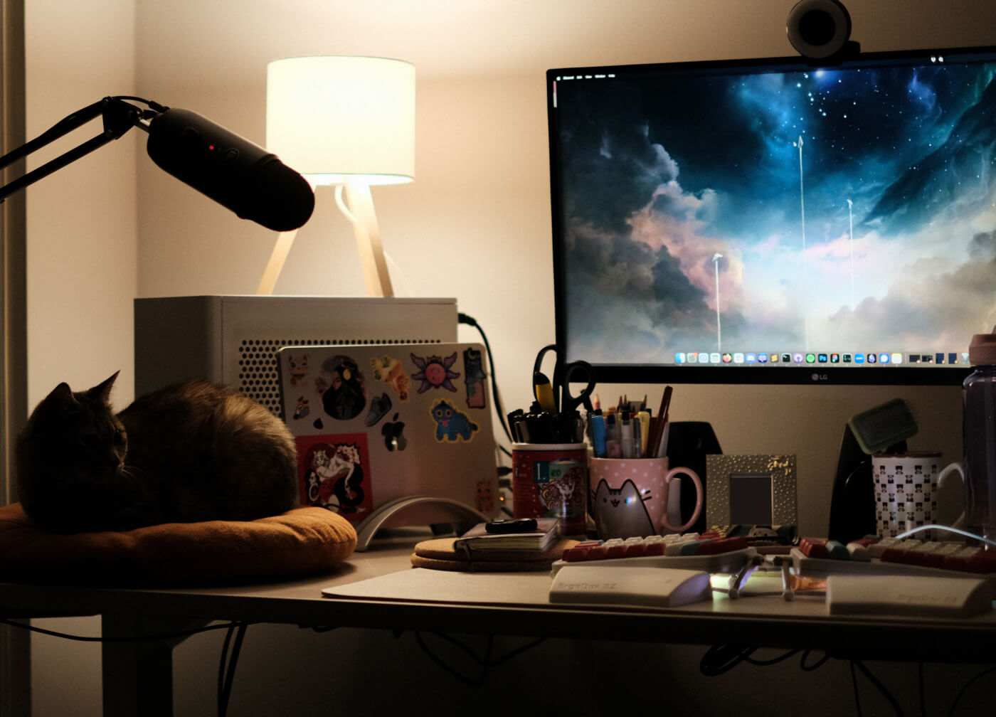 My desk at night, where Sophie sits on a cushion on the side. Everything is mostly in shadow, lit only by a warm lamp; Sophie forms a loaf shape.
